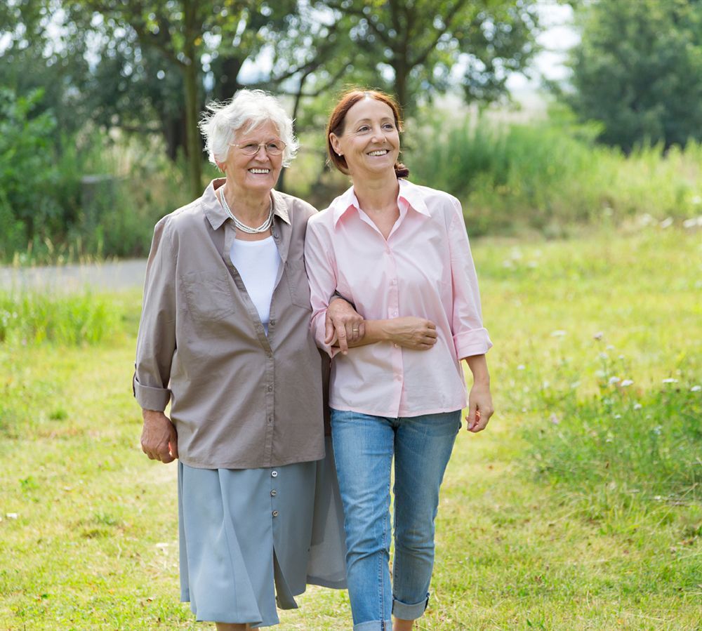 Eine Pflegekraft geht Arm in Arm mit einer Seniorin spazieren als Begleitdienst