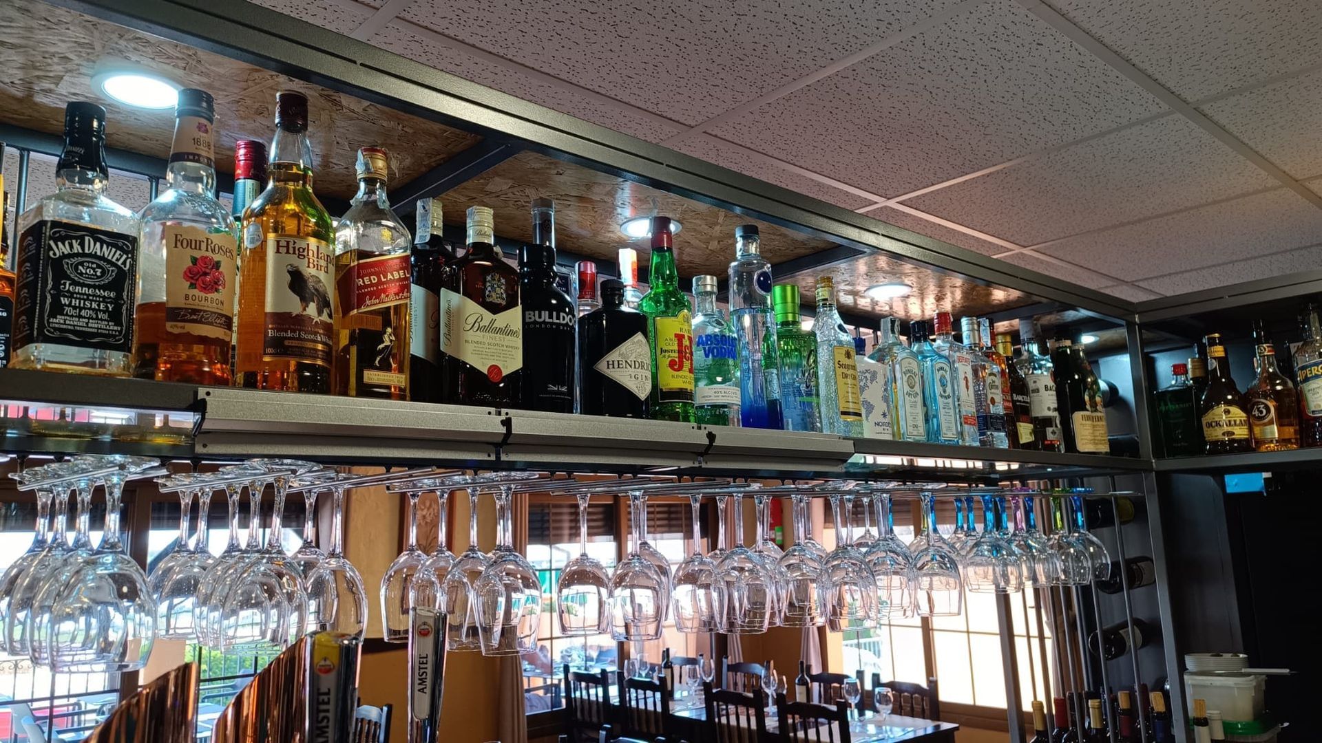 Bar con botellas de licor en estantes, vasos colgando debajo y un comedor a la vista.