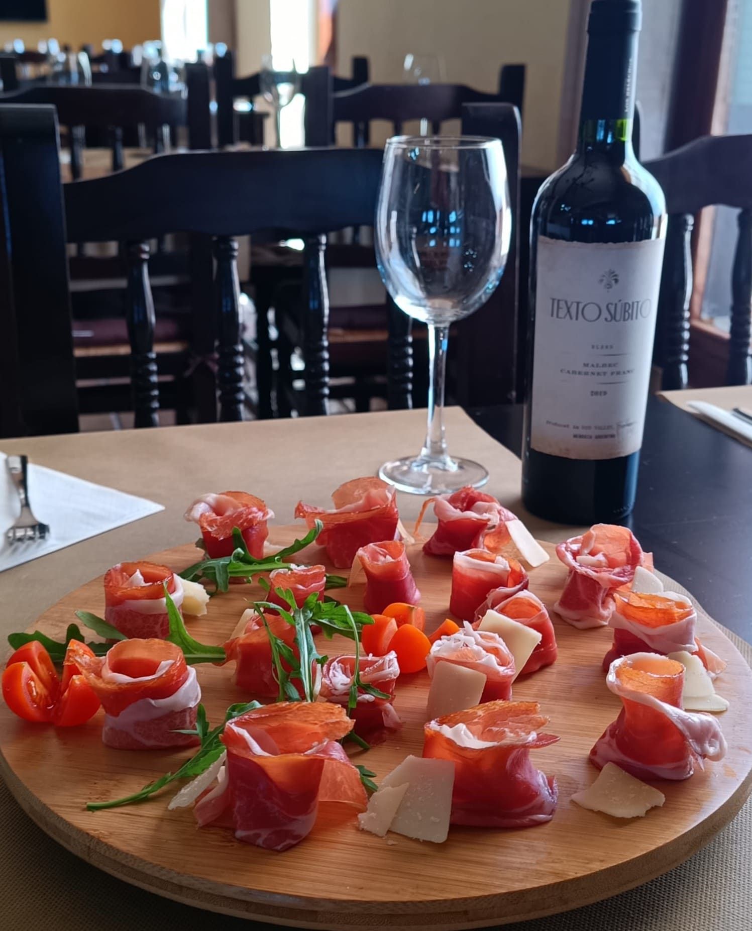 Tabla de madera con tomates envueltos en prosciutto, queso, rúcula, botella de vino, copa y un decorado de restaurante.