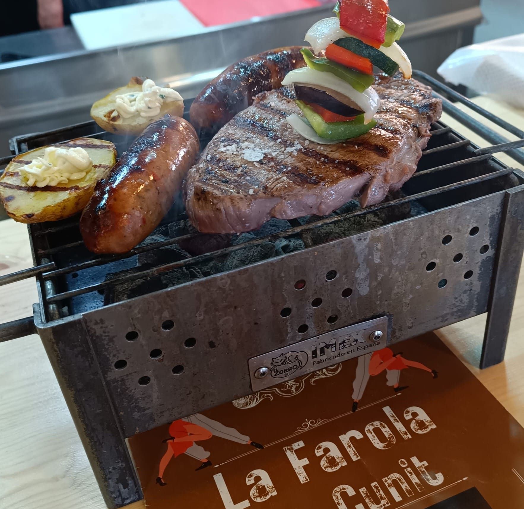 Parrilla con bistec, salchicha, verduras y papas cocinándose. Tonos marrones y grises. Se aprecia la marca 