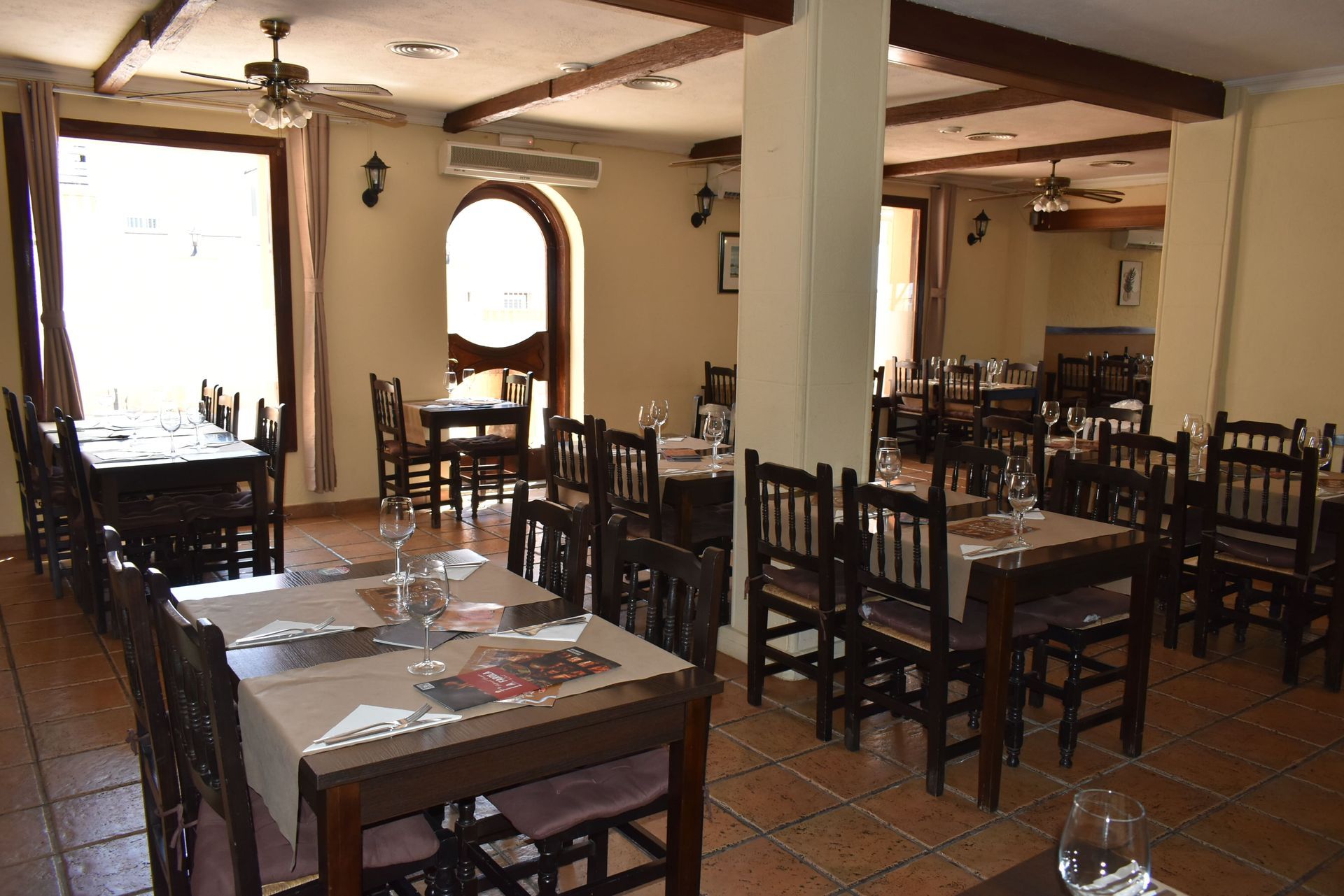 Interior de restaurante con mesas preparadas para comer, sillas marrones, paredes beige y luz natural gracias a la ventana.