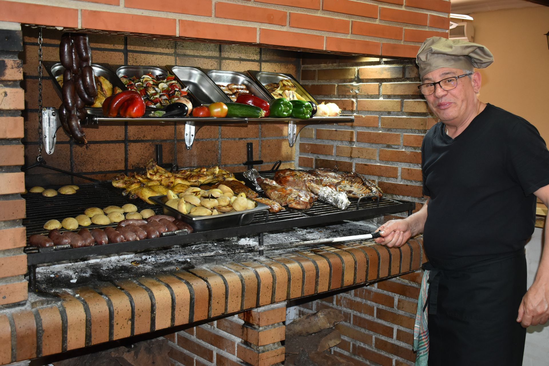 Un chef prepara comida a la parrilla en una barbacoa de ladrillo. Lleva gorro y delantal de chef. Hay varios alimentos cocinados sobre la parrilla y la rejilla.