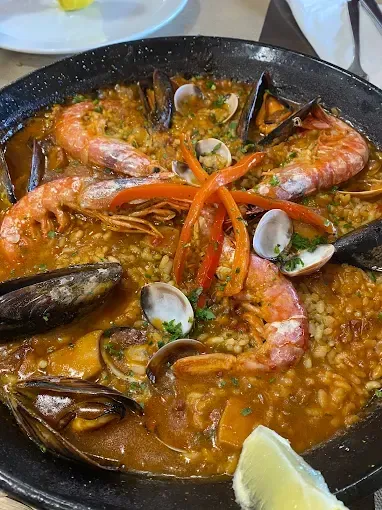 Paella con gambas, mejillones y almejas en una paellera negra grande, con una rodaja de limón al lado.