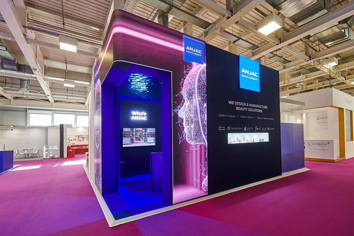 Un stand moderne pour un salon professionnel, avec un logo numérique bleu lumineux, une identité visuelle bleue et une moquette rose sur le sol d'un hall d'exposition.
