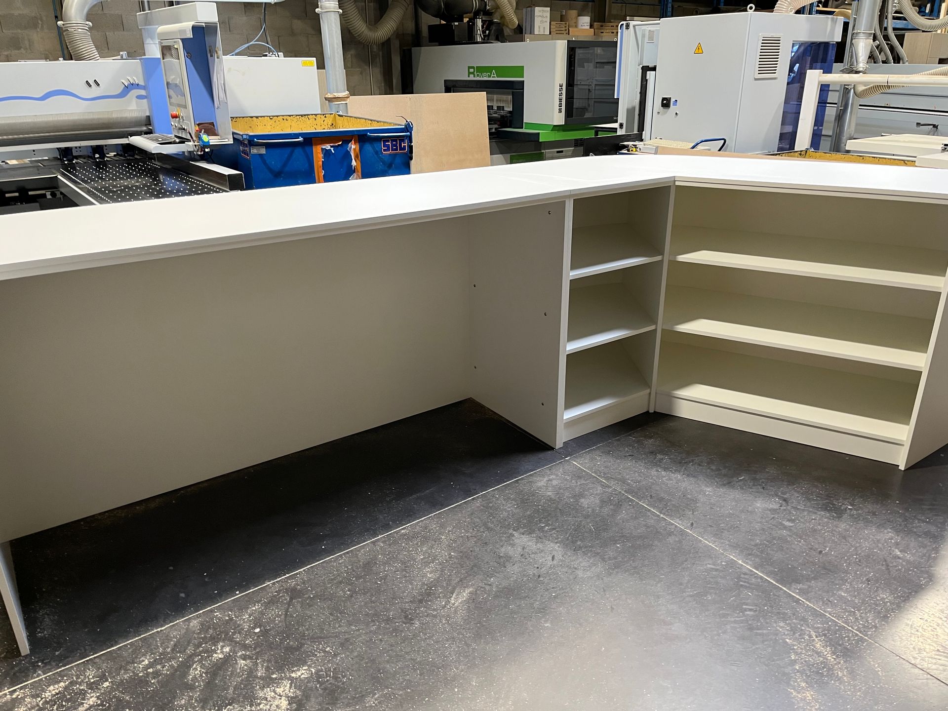Bureau blanc en forme de L avec étagères intégrées, dans un atelier avec des machines en arrière-plan.