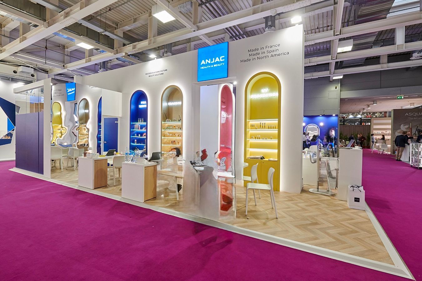 Un stand d'exposition présentant des niches cintrées bleues et jaunes, des comptoirs blancs et des chaises sur un sol en bois.