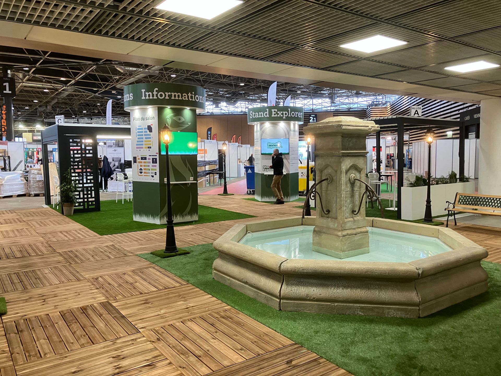 Un salon professionnel intérieur avec une fontaine, du gazon synthétique et des stands présentant des produits d'aménagement paysager.