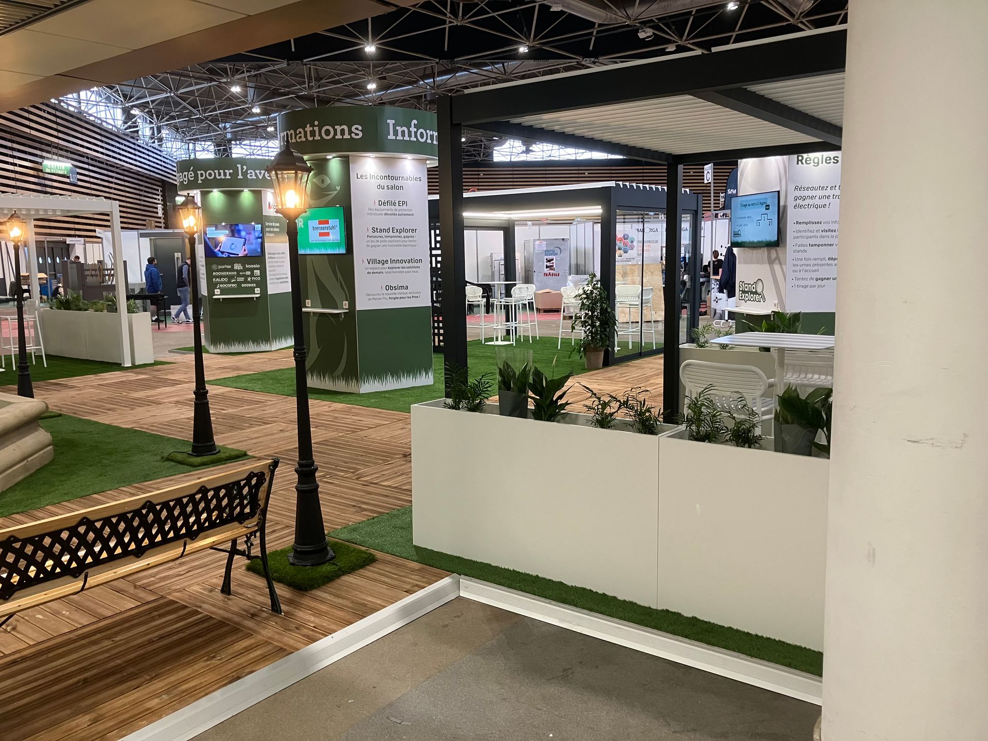 Stand d'exposition avec présentation extérieure, comprenant aménagement paysager, signalétique et sièges.