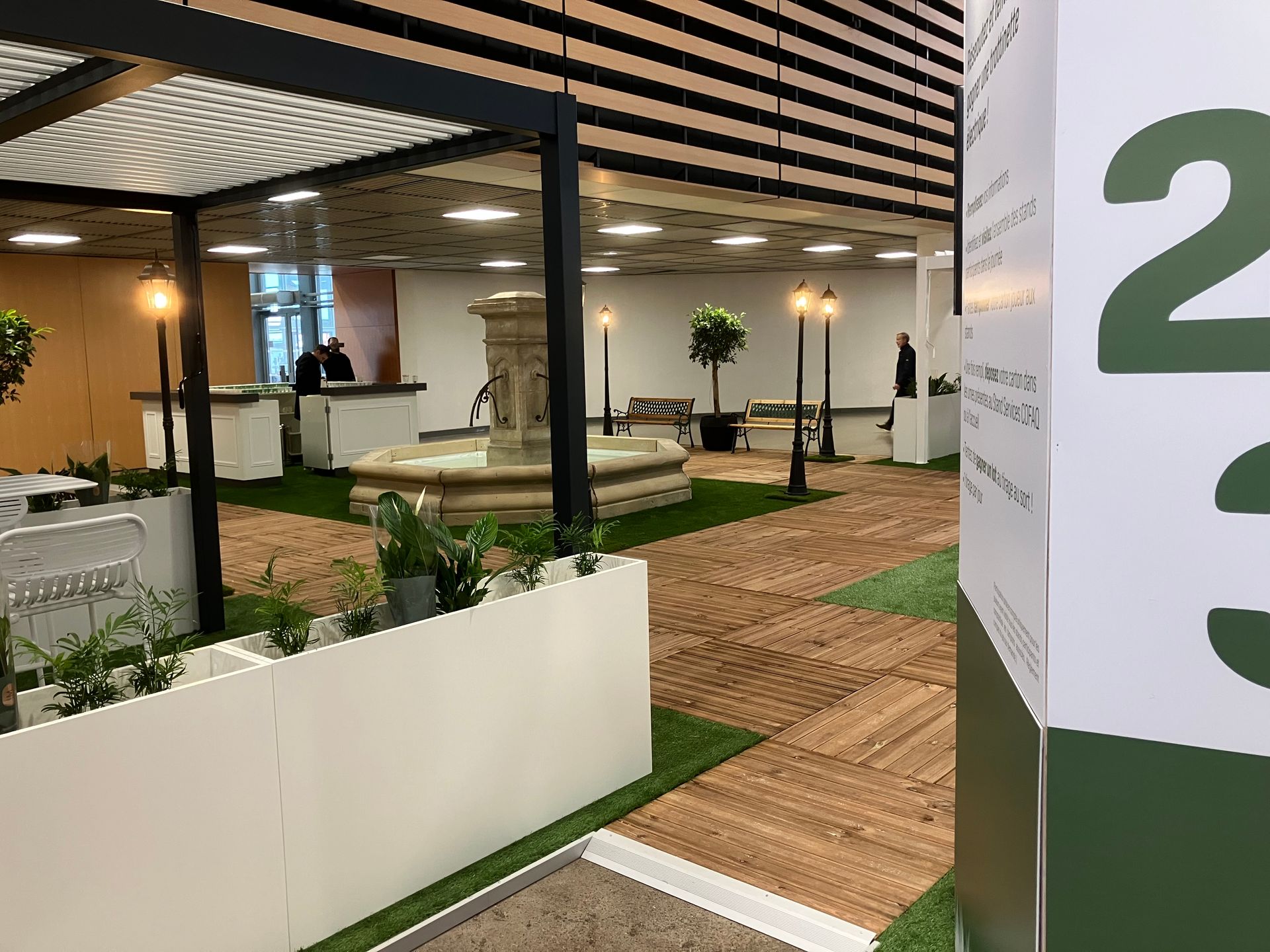 Espace intérieur avec fontaine, verdure et allées en bois. Un panneau portant le numéro 2 se trouve au premier plan.