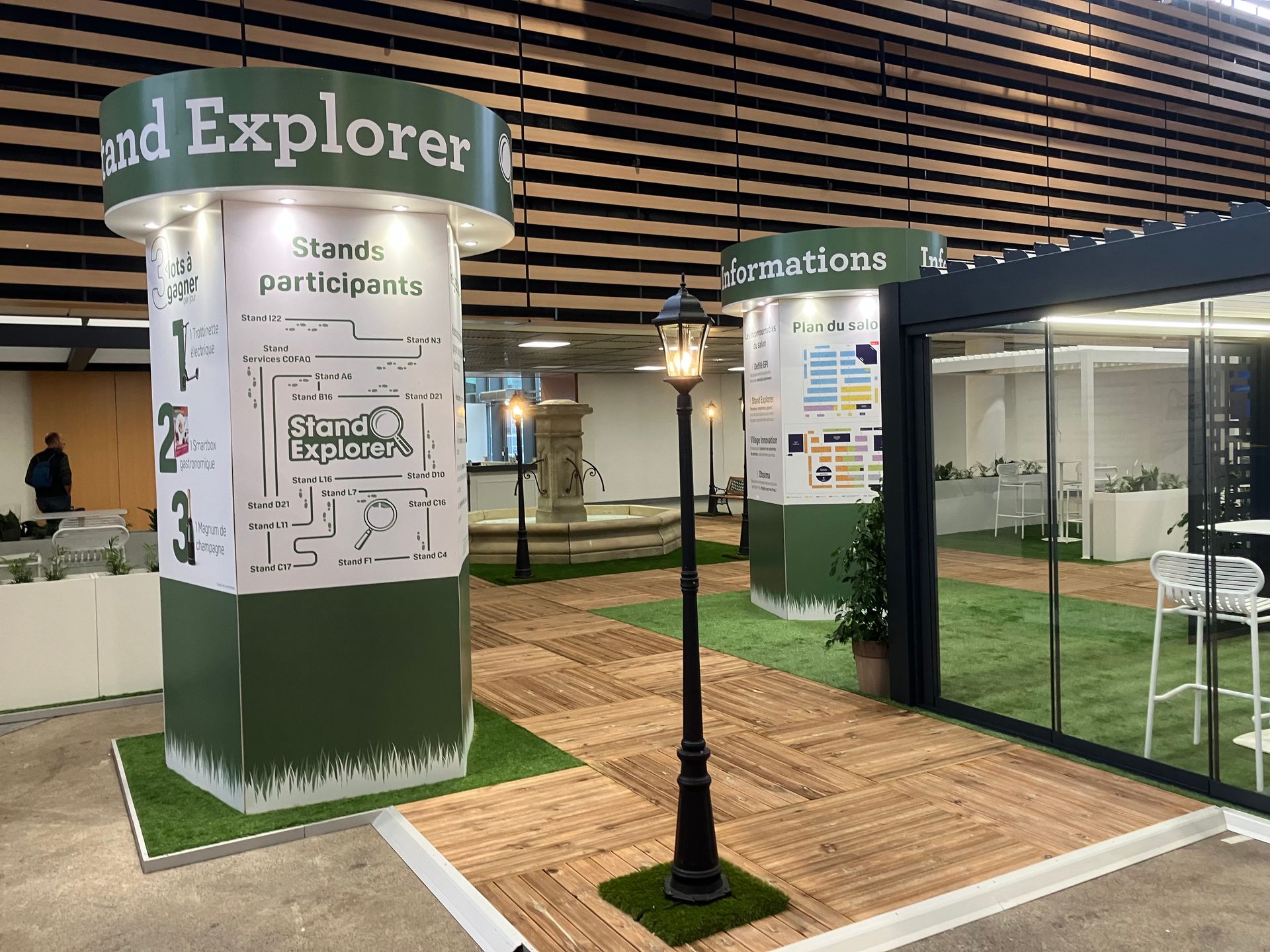 Stand d'exposition avec touches de vert et gazon artificiel. Des colonnes présentent textes et graphiques. Un lampadaire se dresse sur un chemin de bois.