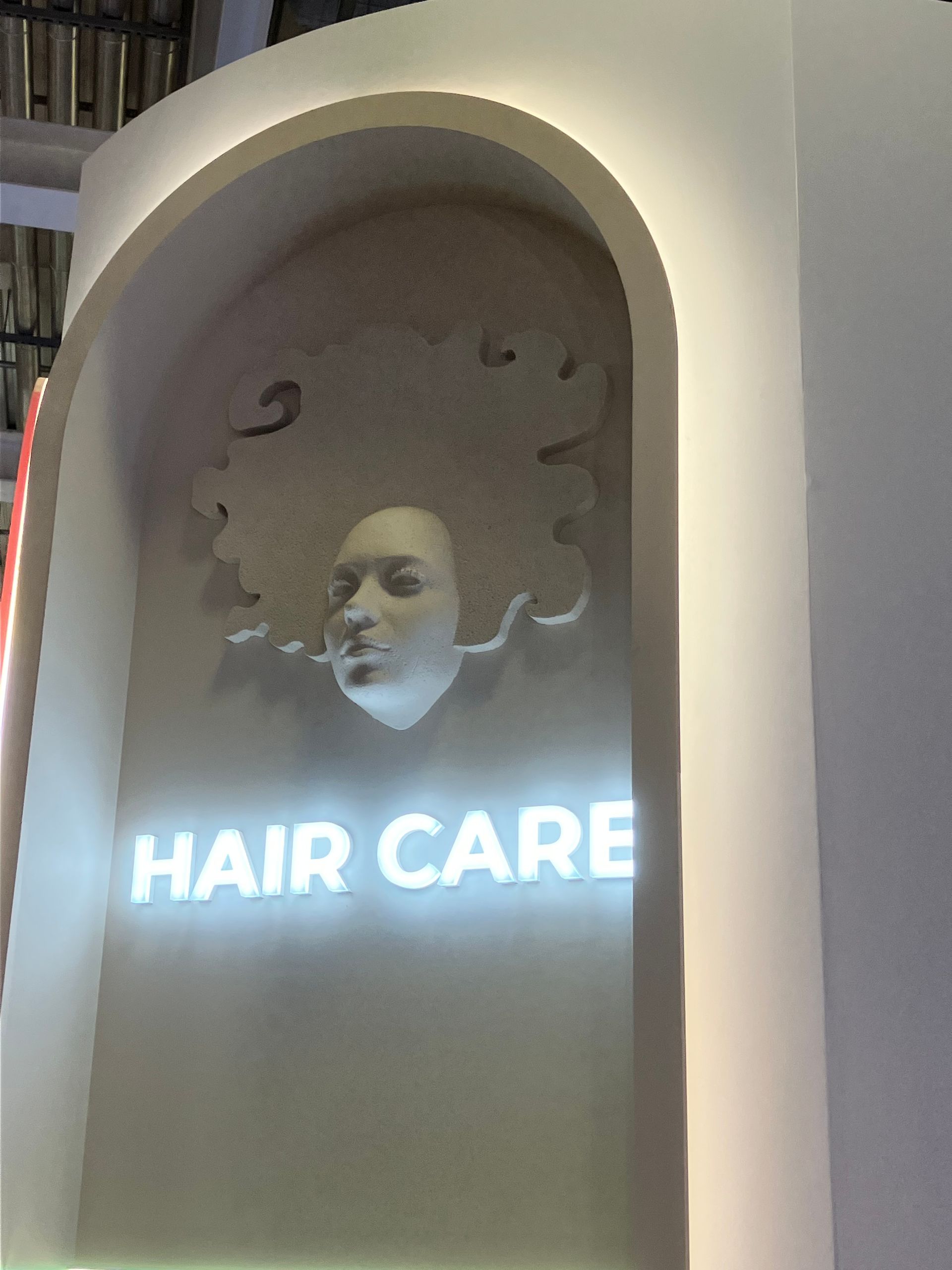 Une enseigne blanche en forme d'arche portant l'inscription SOINS CAPILLAIRES en lettres rétroéclairées sous un visage sculpté en relief.