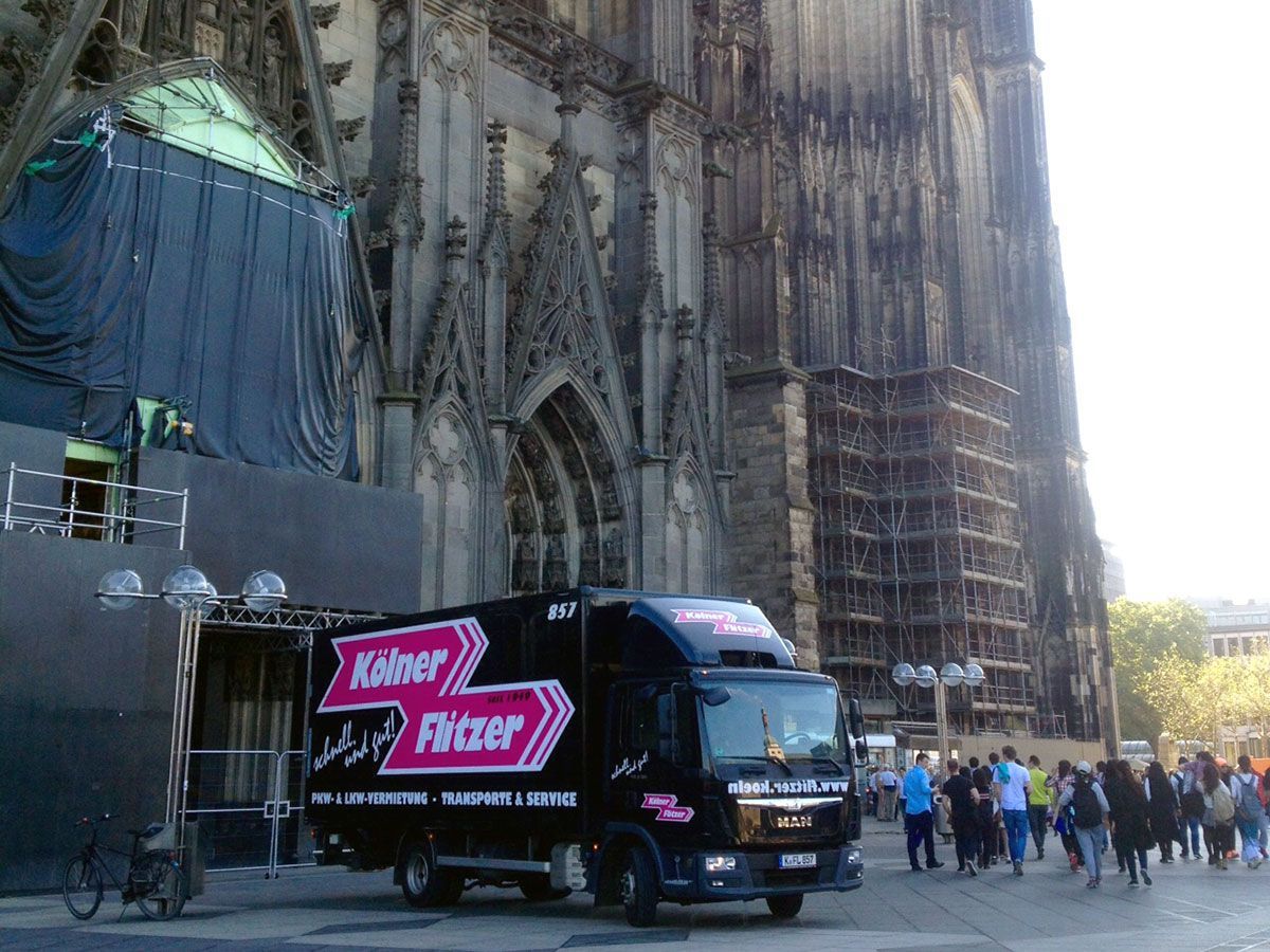 Ein schwarzer LKW mit Kölner Flitzer auf der Seite steht vor einem großen Gebäude