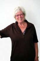 Eine ältere Frau mit Brille und braunem Hemd steht vor einer weißen Wand.