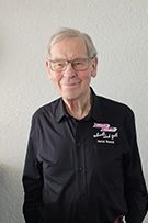 Ein Mann mit Brille und kariertem Hemd steht vor einer weißen Wand.