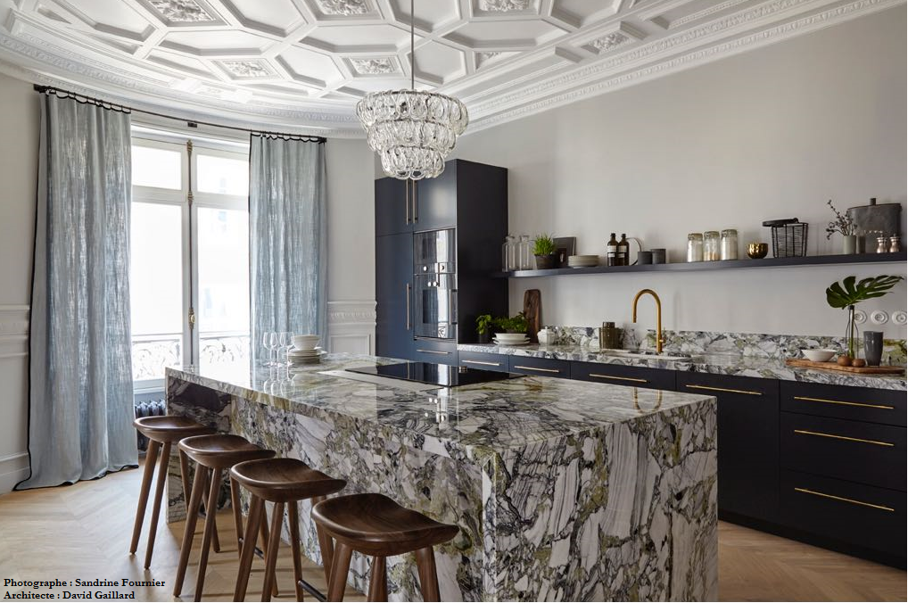 Cuisine luxueuse : îlot en marbre, placards foncés, plafond orné, lustre, tabourets.