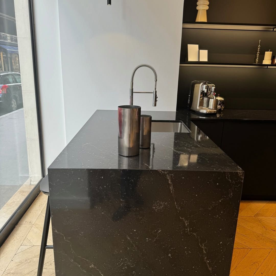 Plan de travail noir avec évier, robinet et récipients. Placards et étagères noirs avec machine à café.