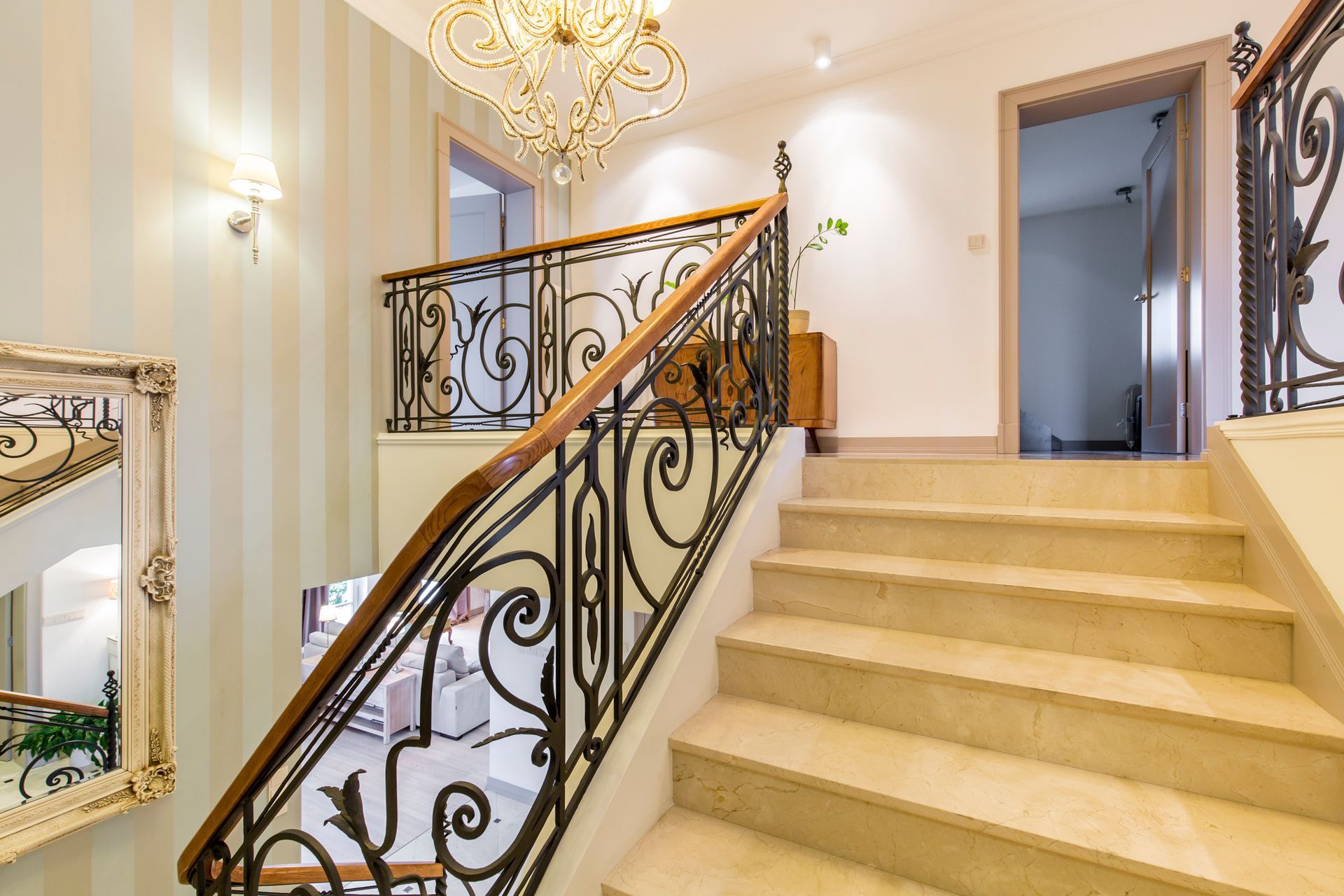 Escalier avec rampe en fer forgé ouvragée, marches couleur crème et lustre décoratif.
