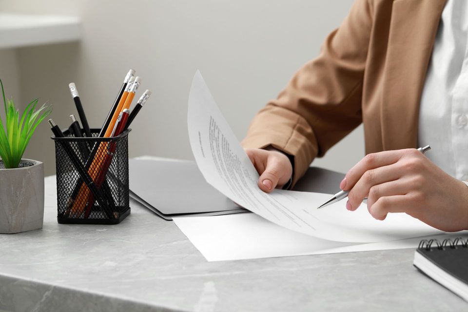 Une personne relit des documents avec un stylo, un bureau avec une plante, des crayons et un carnet.