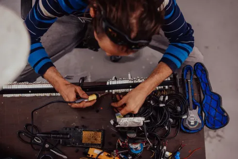 Persona soldando dispositivos electrónicos en un banco de trabajo con guitarra y cables.