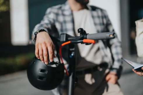 Persona apoyada en un patinete eléctrico, sujetando un casco. Escenario al aire libre.