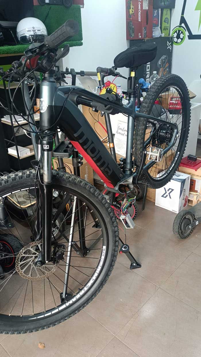 Bicicleta de montaña negra en el interior, junto a otra bicicleta, con un cargador o batería roja acoplada al cuadro.