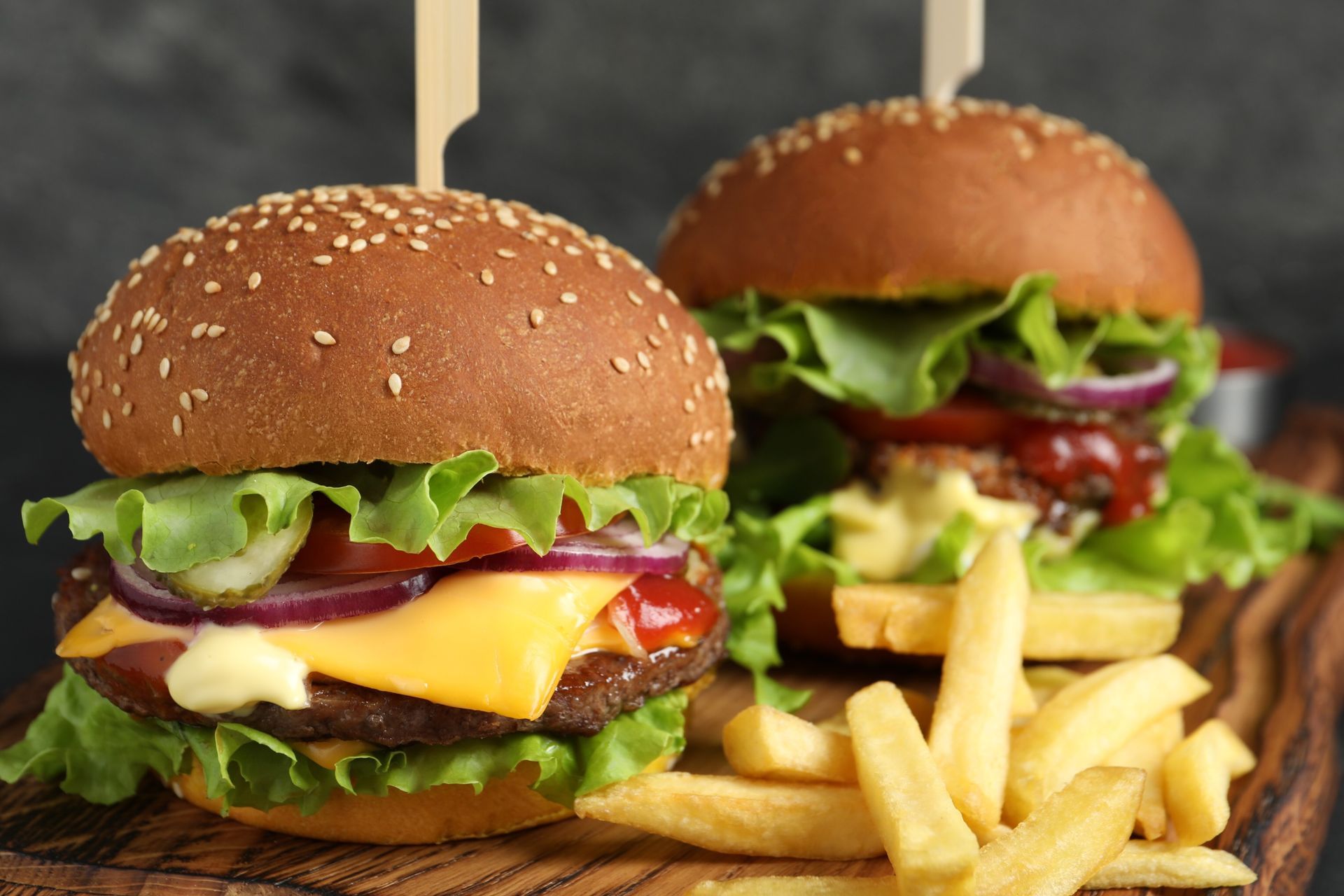 Dos hamburguesas con panecillos de sésamo, queso, lechuga, tomate y cebolla roja; papas fritas sobre una tabla de madera.