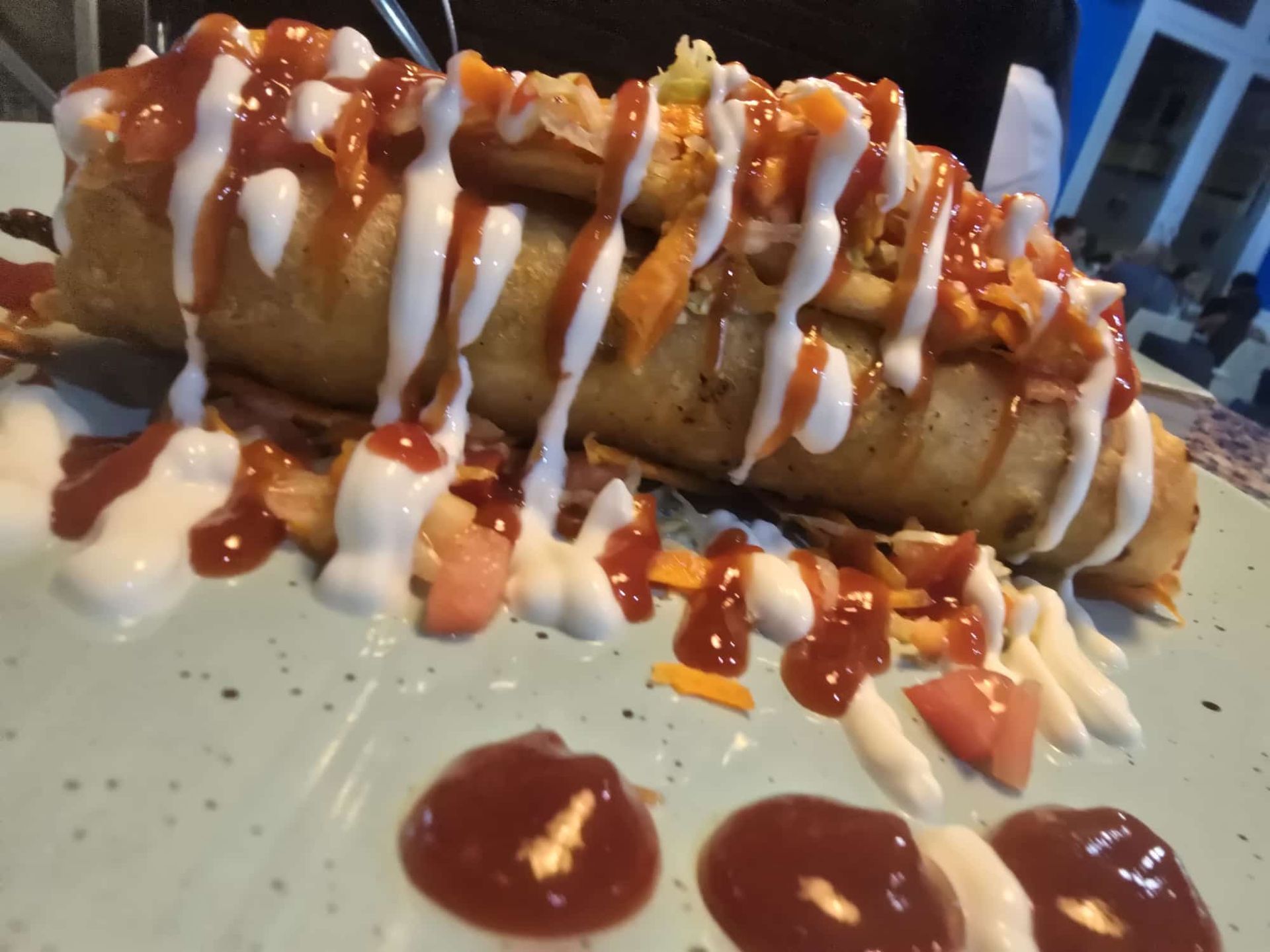 Burrito grande, frito y cubierto con ketchup, salsa blanca y guarniciones en un plato azul moteado.