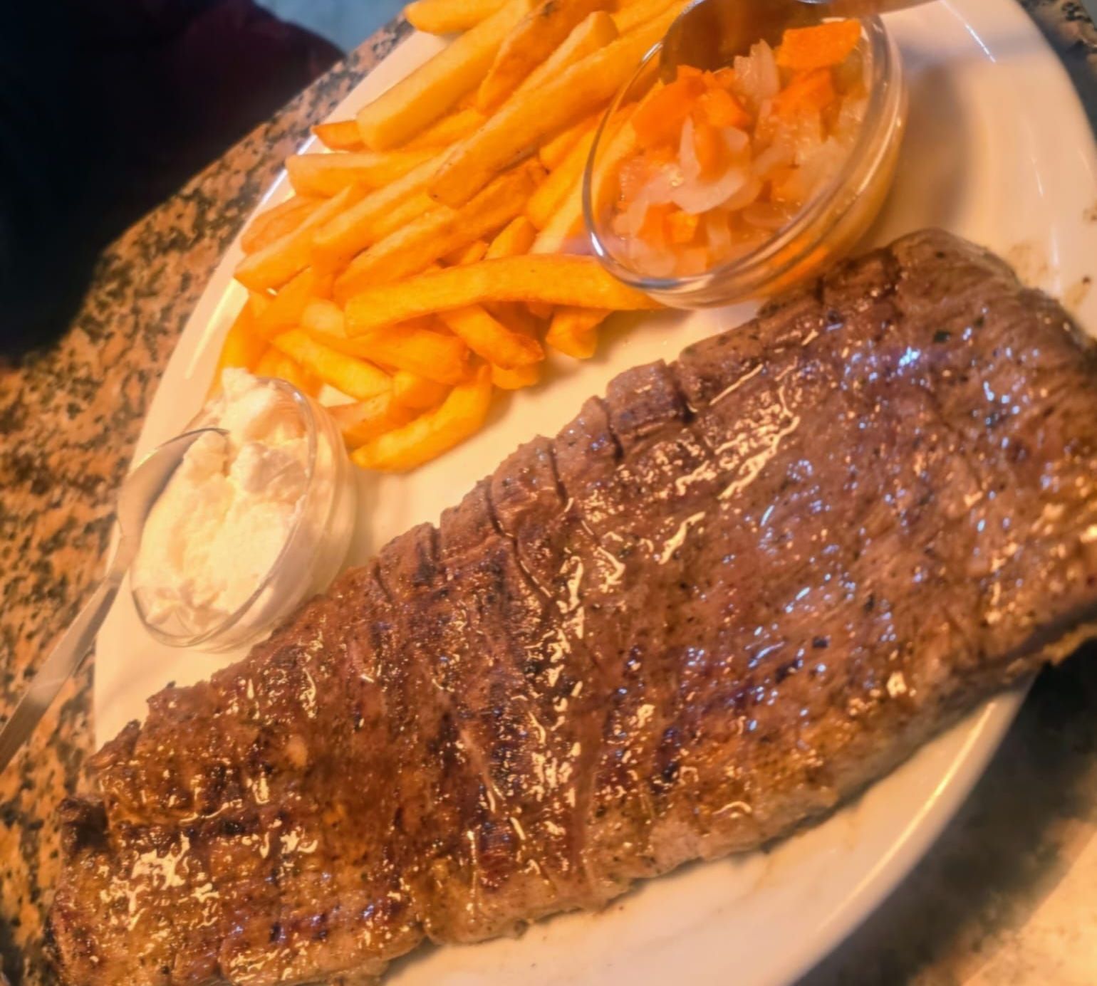 Bistec a la parrilla con papas fritas, condimentos y salsa en un plato blanco.