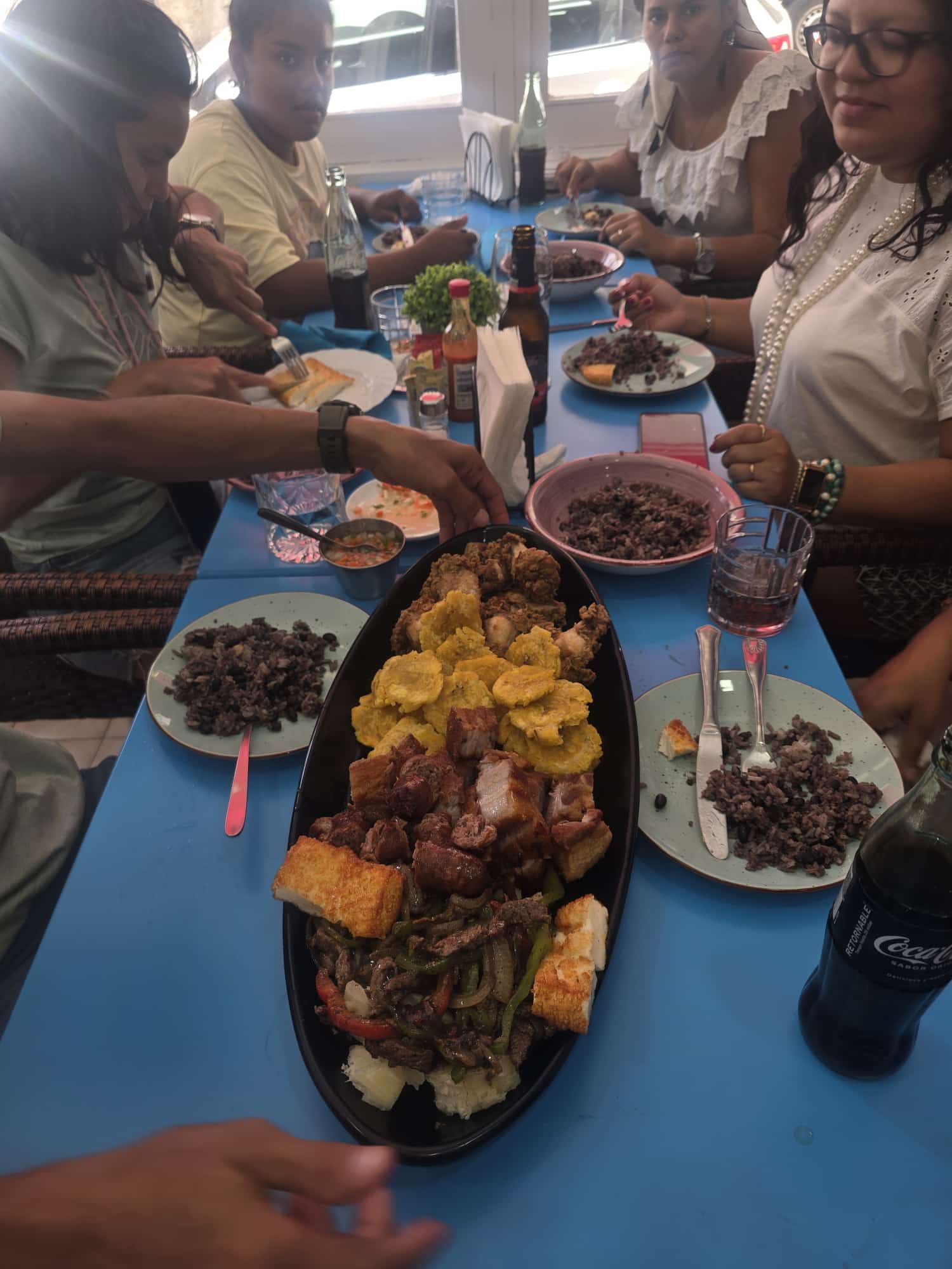 Personas sentadas en una mesa azul, compartiendo un plato de comida: plátanos fritos, cerdo, frijoles y otros artículos.