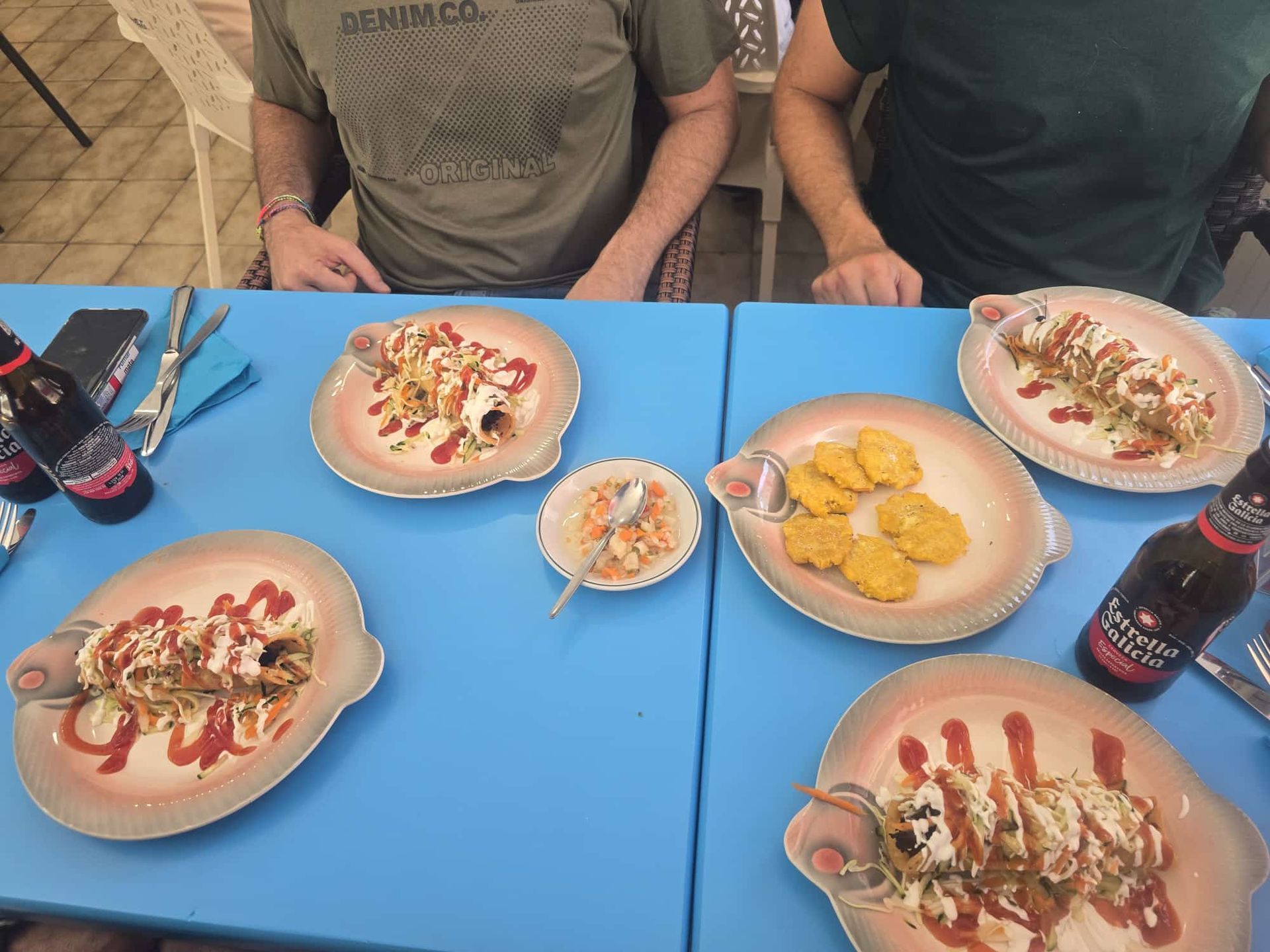 Dos personas en una mesa azul con platos de comida (wraps, nuggets), un tazón pequeño y botellas de cerveza.