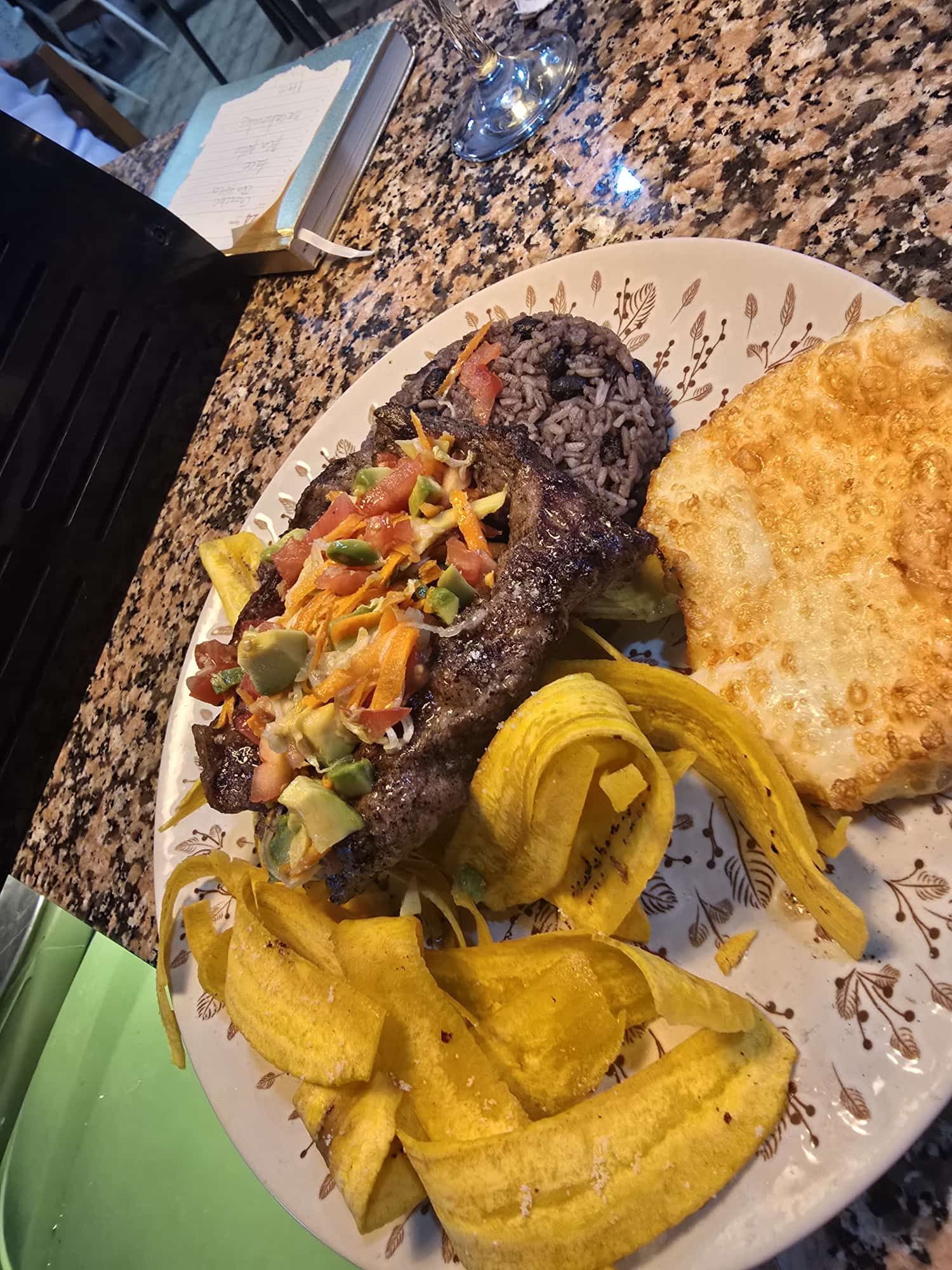 Plato de comida: bistec cubierto con aguacate y salsa, frijoles negros, plátanos fritos y huevo frito.