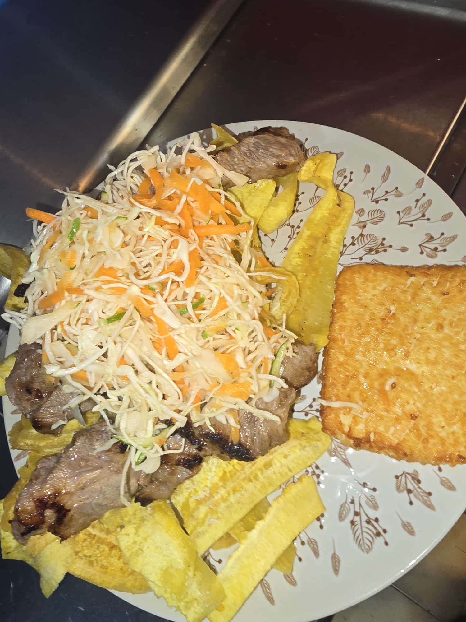 Plato con carne a la parrilla, papas fritas de plátano amarillo, ensalada de col rallada y una papa hash brown dorada.