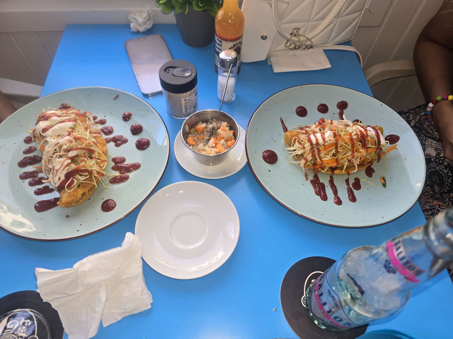Dos platos de comida sobre una mesa azul con condimentos y una bebida.
