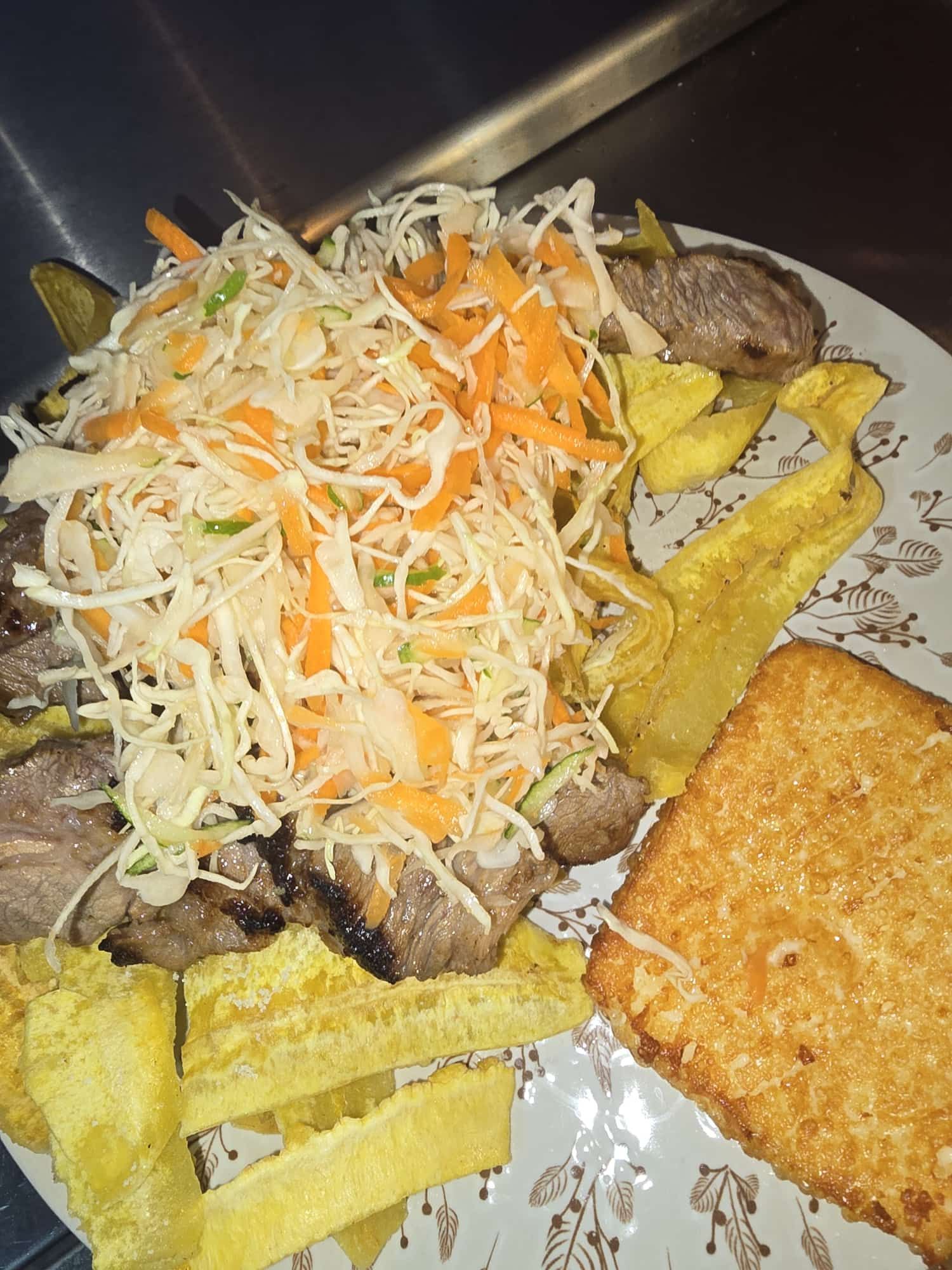 Plato de carne a la parrilla con repollo rallado, zanahoria, plátanos fritos y papa hash brown.