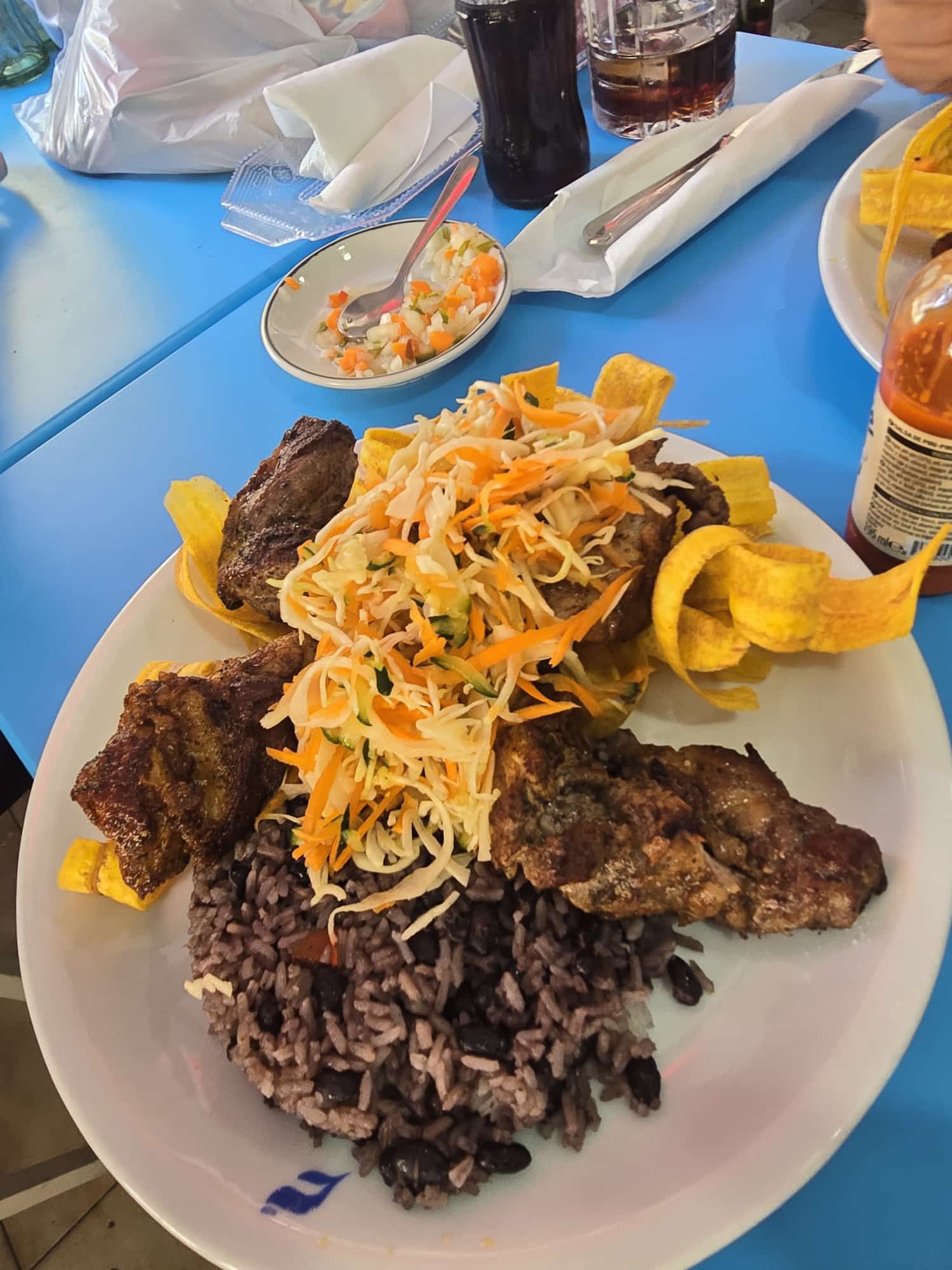Plato de comida: carne a la parrilla, frijoles negros con arroz, chips de plátano y ensalada de col sobre una mesa azul.