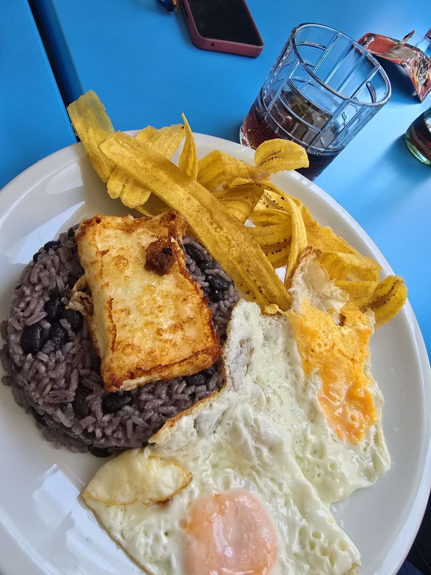 Plato con arroz, frijoles, huevo frito, queso frito y plátanos. Mesa azul y bebida.