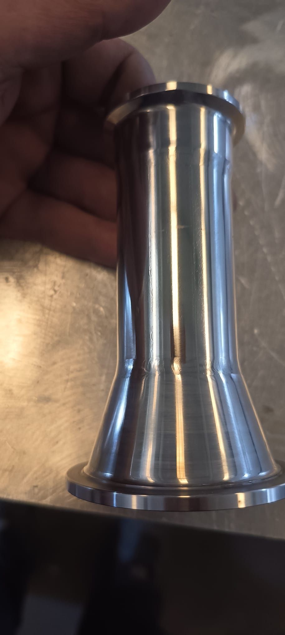Una pieza mecanizada de metal brillante sostenida por la mano. Tiene base y parte superior con bridas y forma cónica.