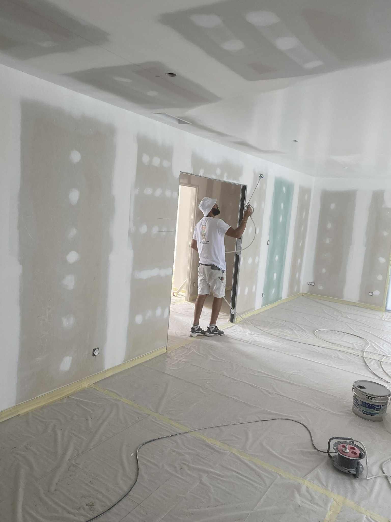 Un homme peint un mur dans une pièce en rénovation ; cloisons sèches non finies, revêtement de sol protecteur.