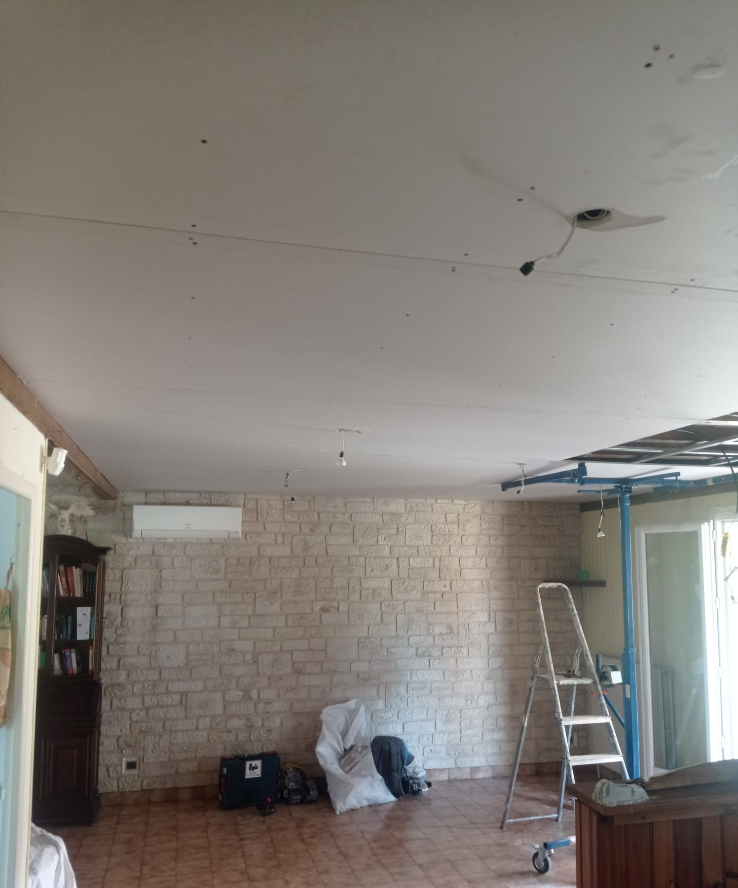 Vue intérieure d'une pièce avec un mur en pierre et des câbles électriques apparents au plafond ; une échelle est appuyée contre le mur.