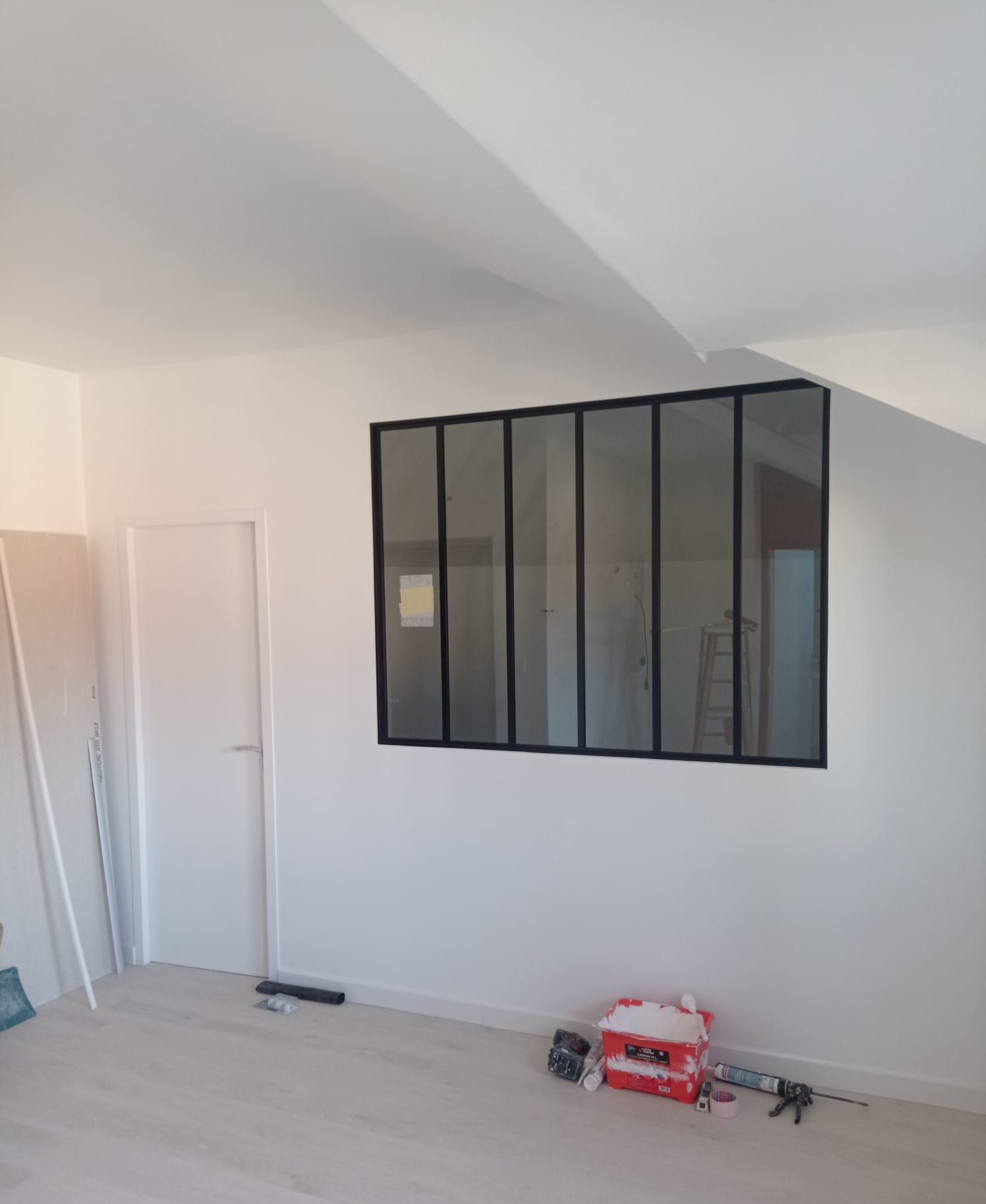 Pièce intérieure avec une grande fenêtre à cadre noir. On aperçoit une porte blanche fermée et des outils au sol.