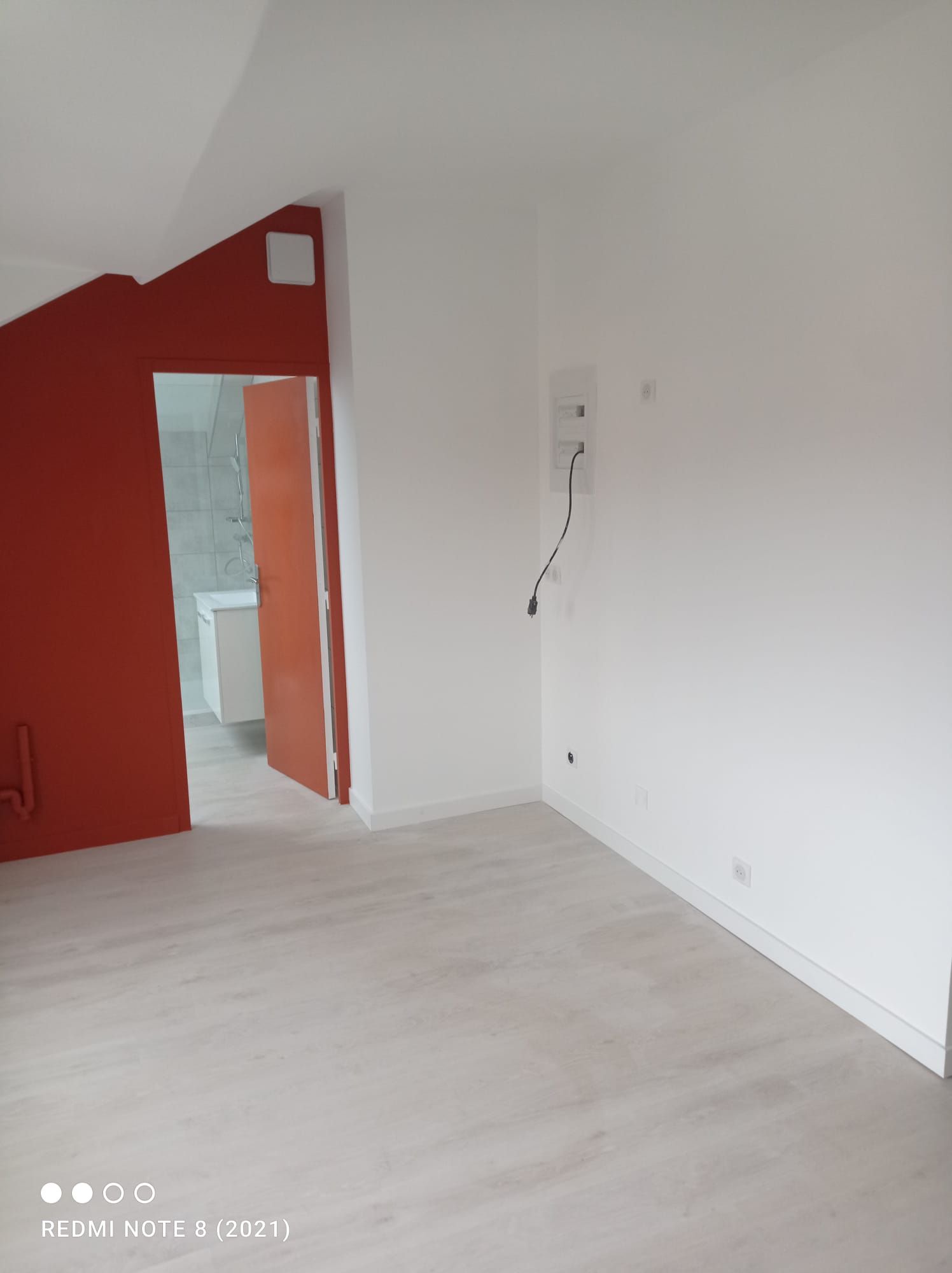 Pièce vide aux murs blancs, parquet clair et encadrement de porte orange donnant sur une autre pièce.