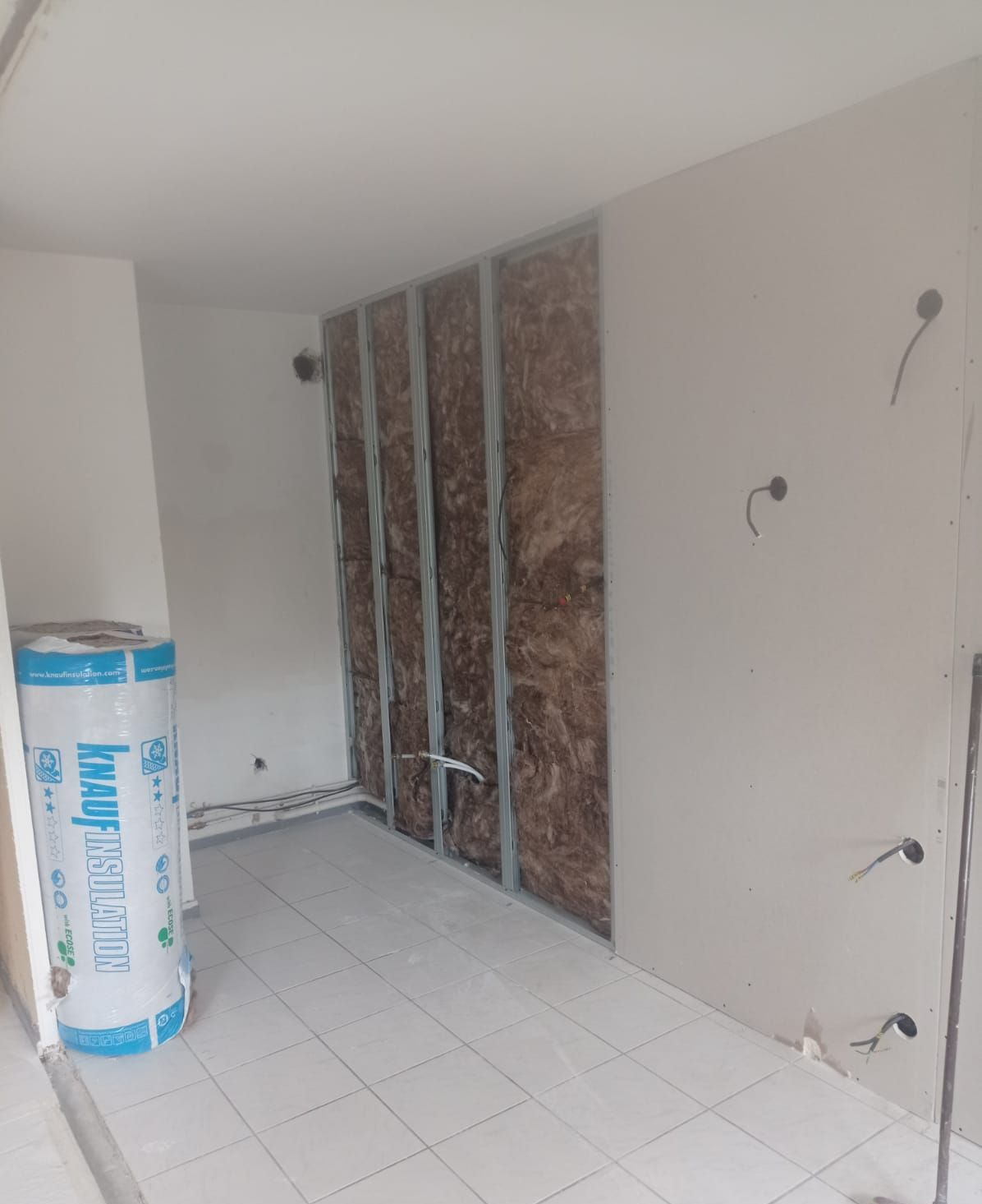 Construction intérieure : ossature murale avec isolation, câblage électrique, plaques de plâtre, carrelage au sol et rouleau d’isolant.