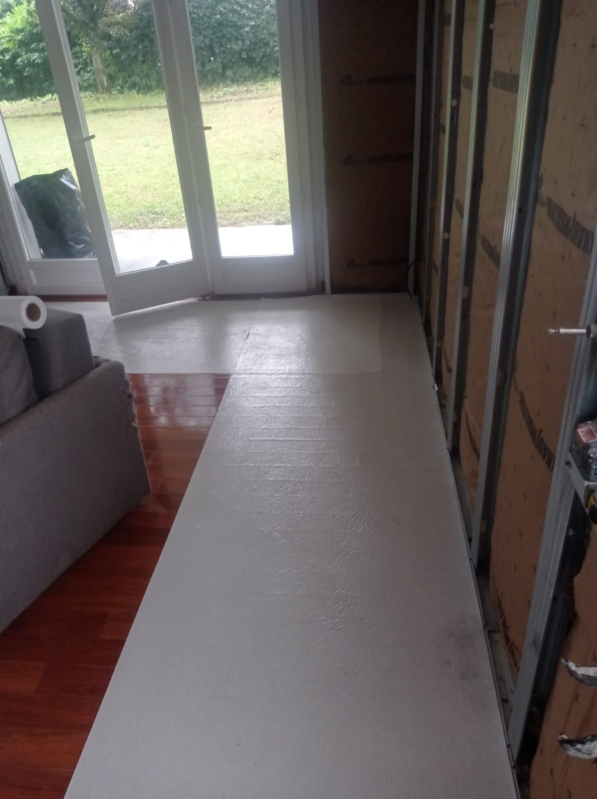 Une pièce avec un sol clair fraîchement peint et des montants muraux apparents, donnant sur des portes vitrées et une vue sur l'extérieur.