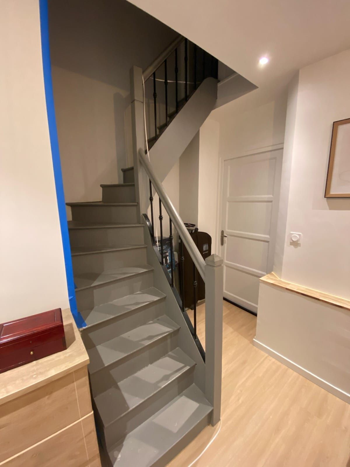 Escalier gris, rampe en bois, menant à un palier. Murs blancs, parquet et porte blanche fermée.