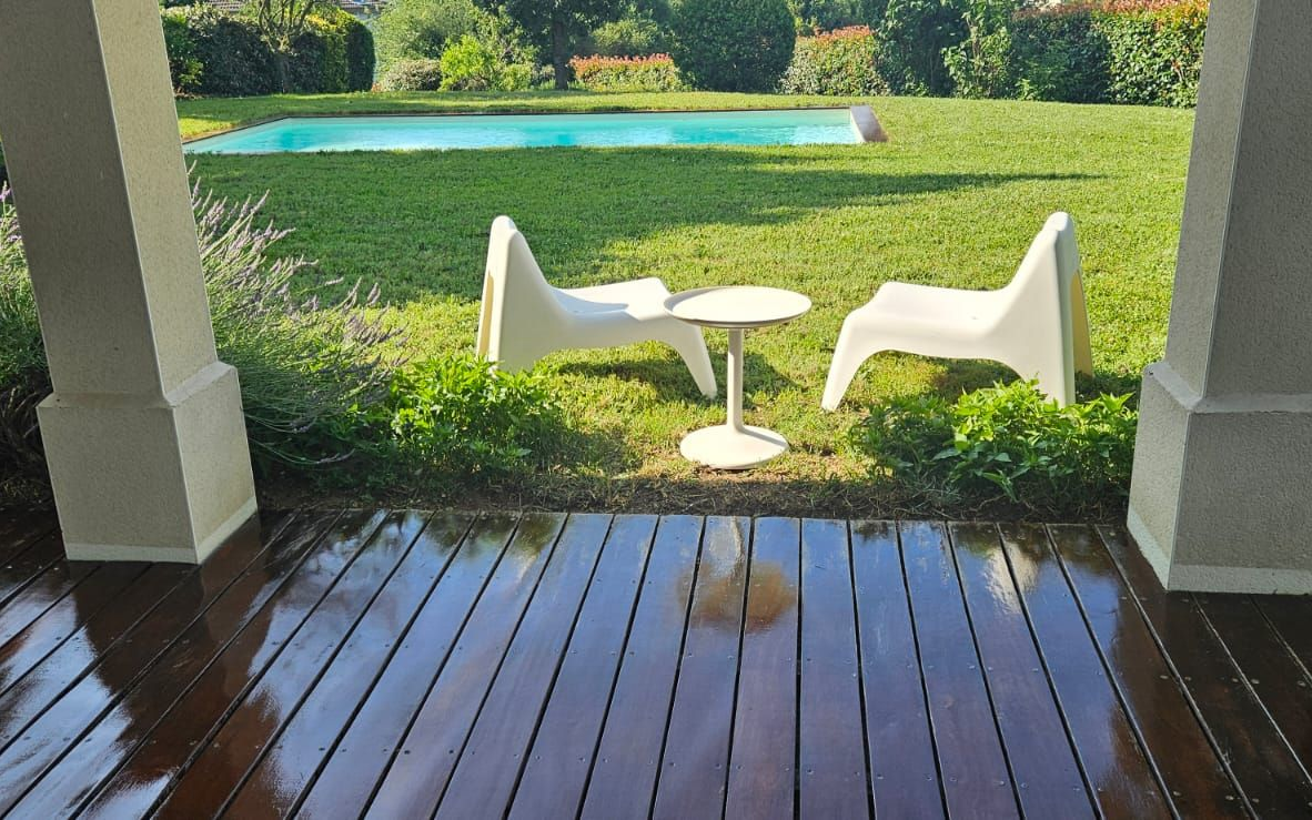 Une terrasse en bois donne sur une pelouse avec piscine, deux chaises blanches et une petite table.