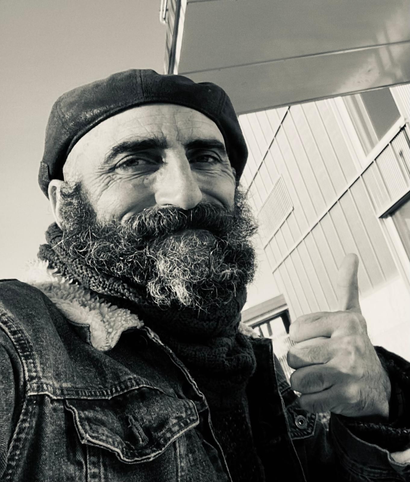 Un hombre con barba larga y boina levanta el pulgar. Al aire libre, cerca de un edificio.