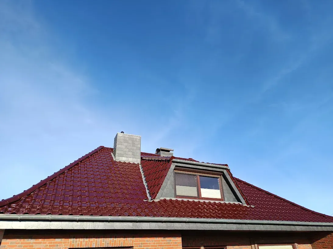Das rote Ziegeldach eines Hauses mit Dachgaube und Schornstein vor blauem Himmel.