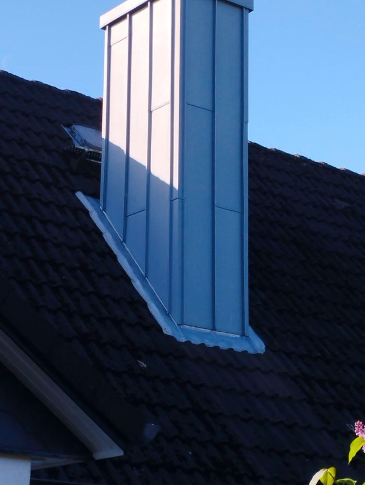 Ein mit Metall verkleideter Schornstein auf einem dunklen Schindeldach vor blauem Himmel.