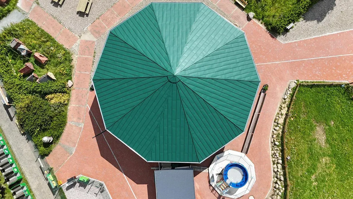 Draufsicht auf ein grünes, achteckiges Gebäude mit Dach auf einem mit Ziegeln gepflasterten Boden neben einer grünen Rasenfläche.
