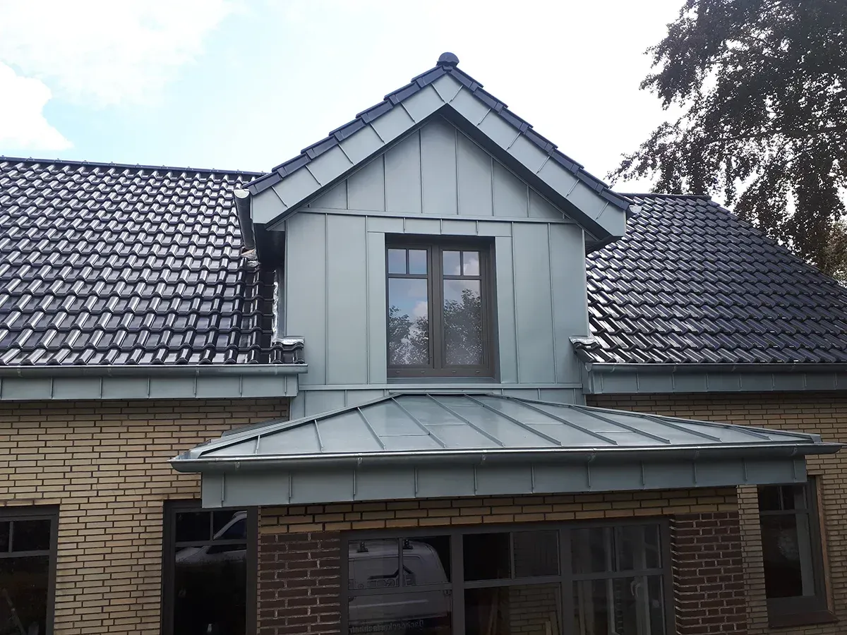Dachgaube mit grünem Metalldach und Backsteinfassade. Schwarzes Ziegeldach. Brauner Fensterrahmen.